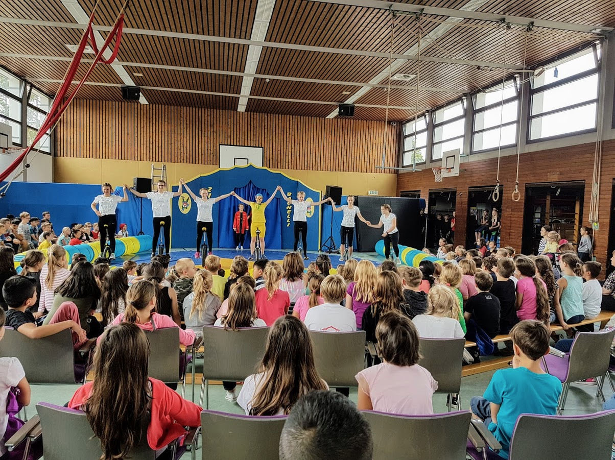 Mehrere Künstler stehen auf einer Zirkus Bühne auf Einrädern vor vielen Kindern die fröhlich zusehen und halten sich an der Hand